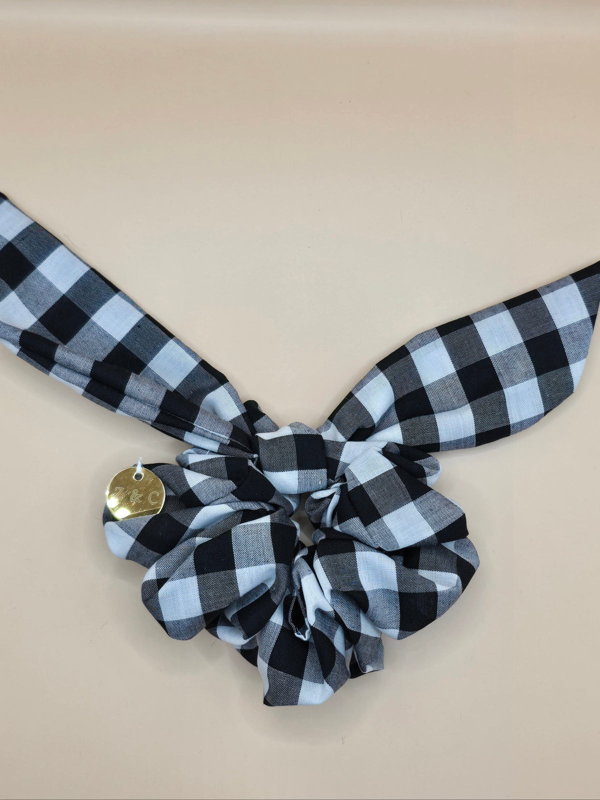 A black and white buffalo check gingham hair scrunchie with a matching tie tail and a small gold button accent, displayed on a solid beige background.