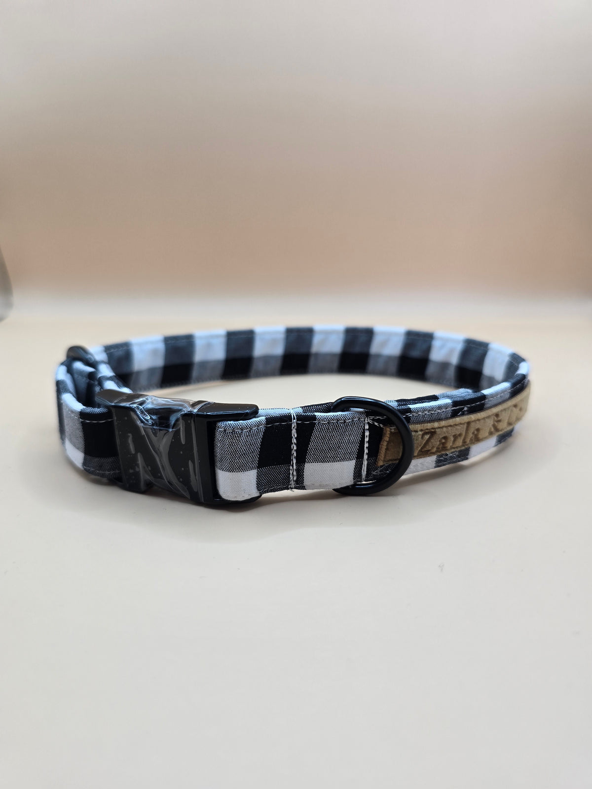 A black and white buffalo plaid dog collar with a black metal buckle and a tan leather tag, shown on a neutral beige background.